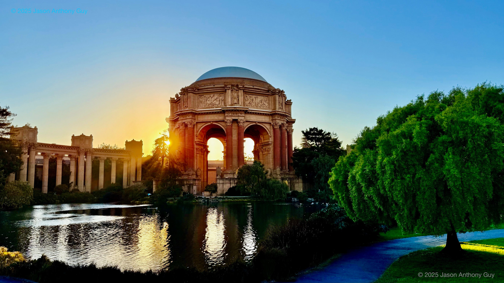 The late afternoon sun peeks out behind the columns of the Palace of Fine Arts, illuminating it from the rear and reflecting off the lake in front. A green tree to the right contrasts with the blue sky and orange glow.