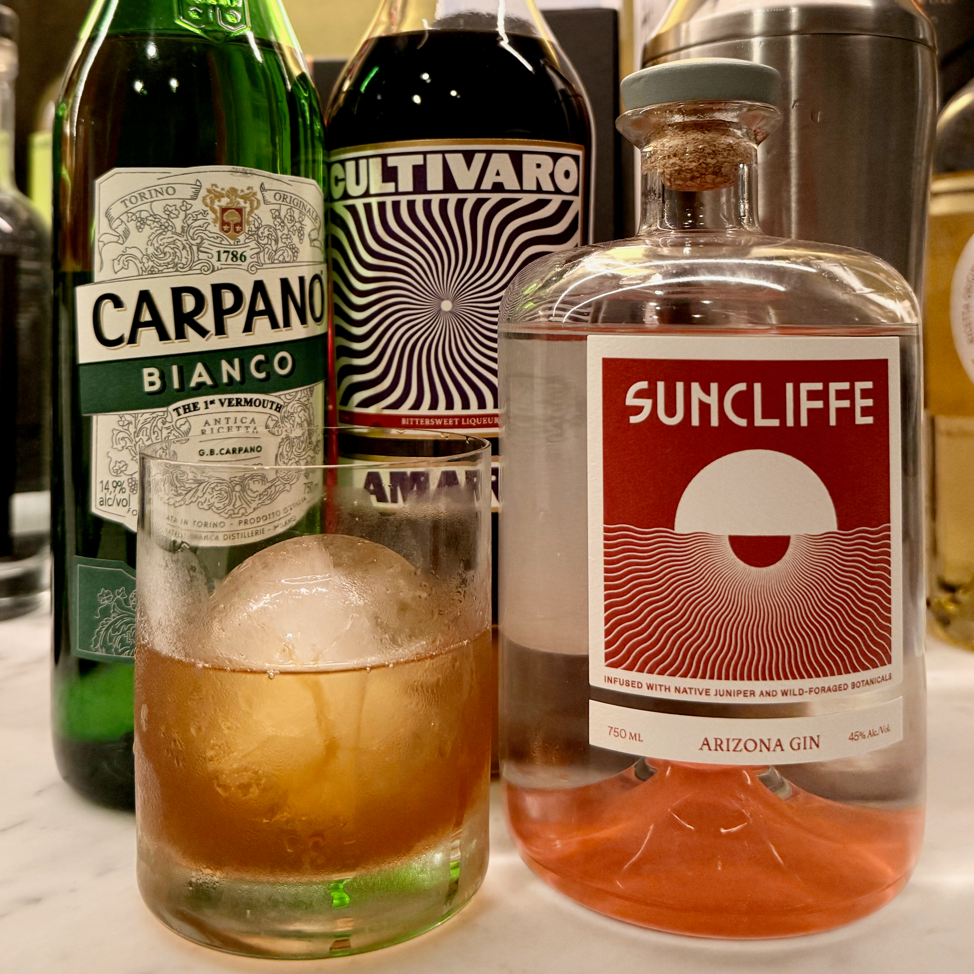 An old fashioned glass filled with a New Age Negroni over a large sphere of ice stands in front of the three ingredients of the cocktail and a metal mixer.