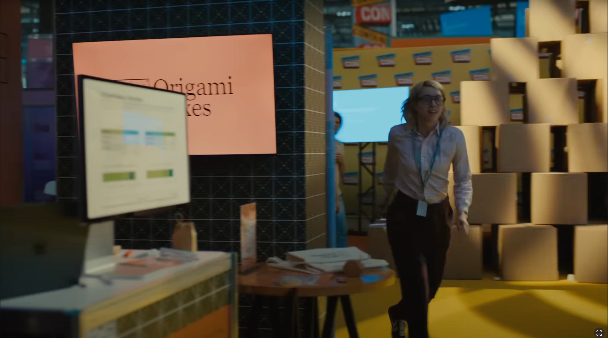 Screenshot from the commercial showing a still-working MacBook and Apple Studio display on the left, with a woman hastily running past it.