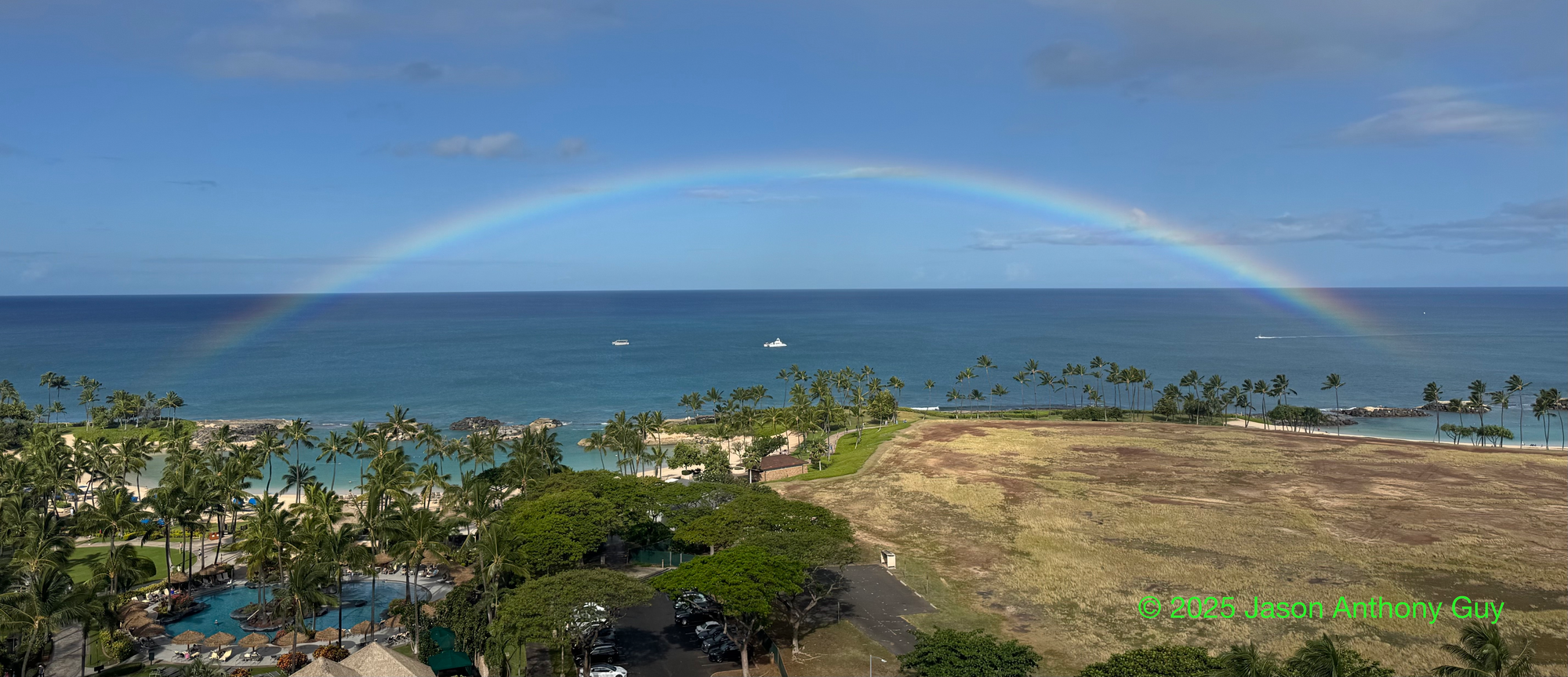 Alt: Photograph of a full arc rainbow off the coast of Oahu. The pale rainbow colors contrast with the bright green of palm trees to the left and the brown-green of empty grassland on the right, with a bright blue ocean and sky as the background.