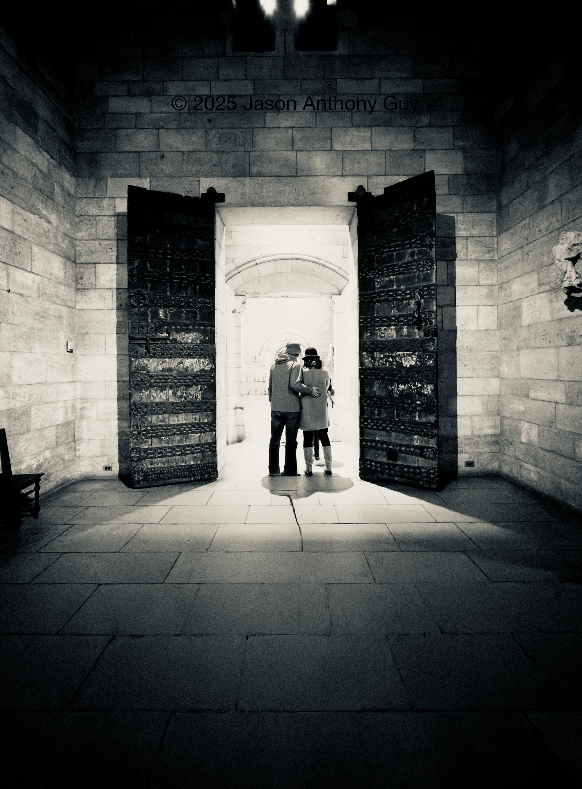 Alt: Black and white photograph of a couple, their backs to us, standing in a doorway with ornate doors, under a bright pool of light  The doors are dark wood or metal, which contrasts with the light colored bricks of the building. The light fades off quickly, leaving the foreground in shadow.