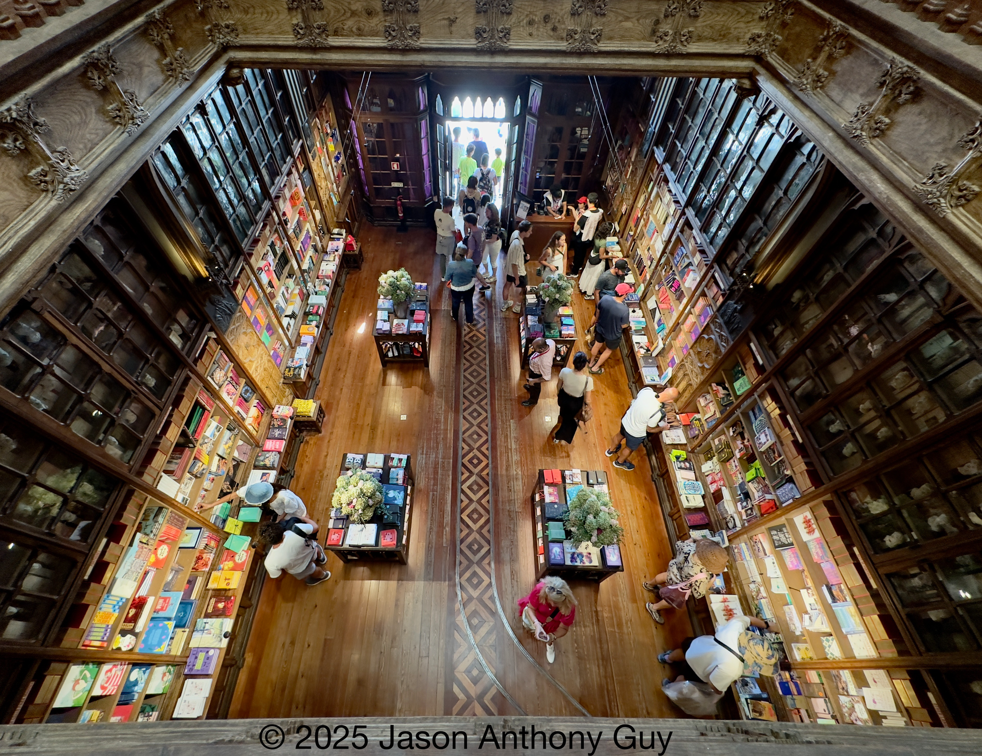 Alt: Photography from above looking down to the lower level through ornate wood railings. The brown wood melds into brown shelves, which morphs into colorful and brightly lit books. People mill about examining books.