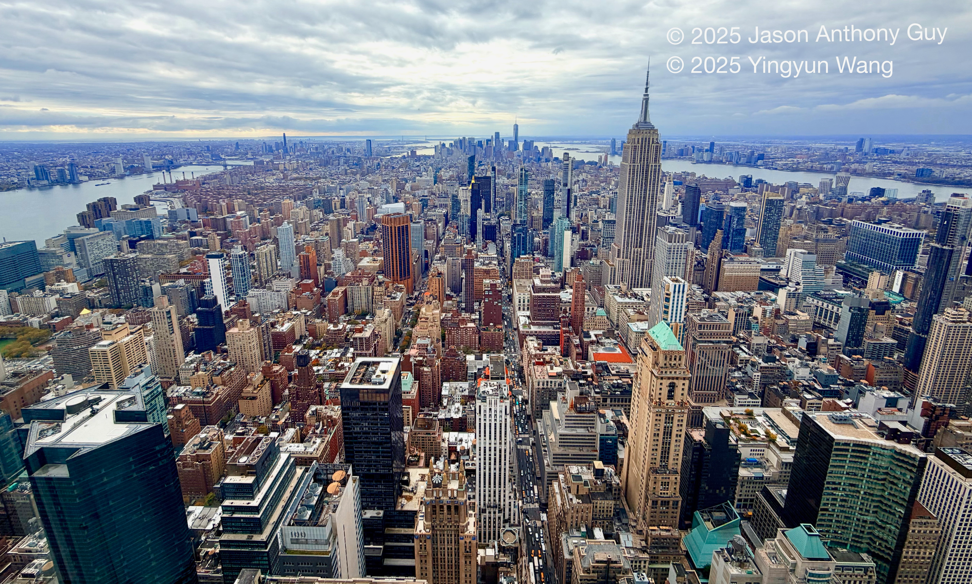 Alt: Photograph of New York City, from midtown looking south, with the East and Hudson rivers flanking the island. The grey-blue skies and river contrast with the dusky browns and tans of the buildings. The image stretches out to a vanishing point in the distance. 