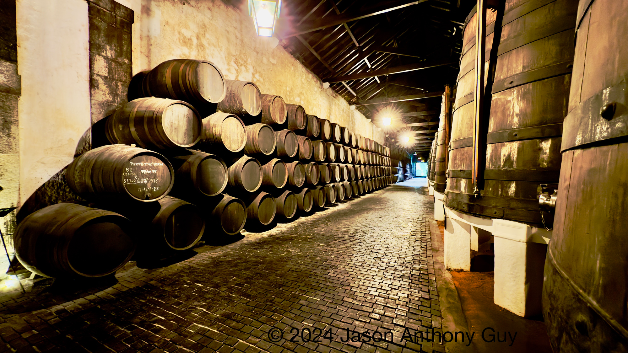 Alt: Photograph of dozens of brown wooden barrels stacked on the left, large wooden vats to the right, with a long brick path leading out a door between them. The image is sepia-toned, evoking age and sentimentality.