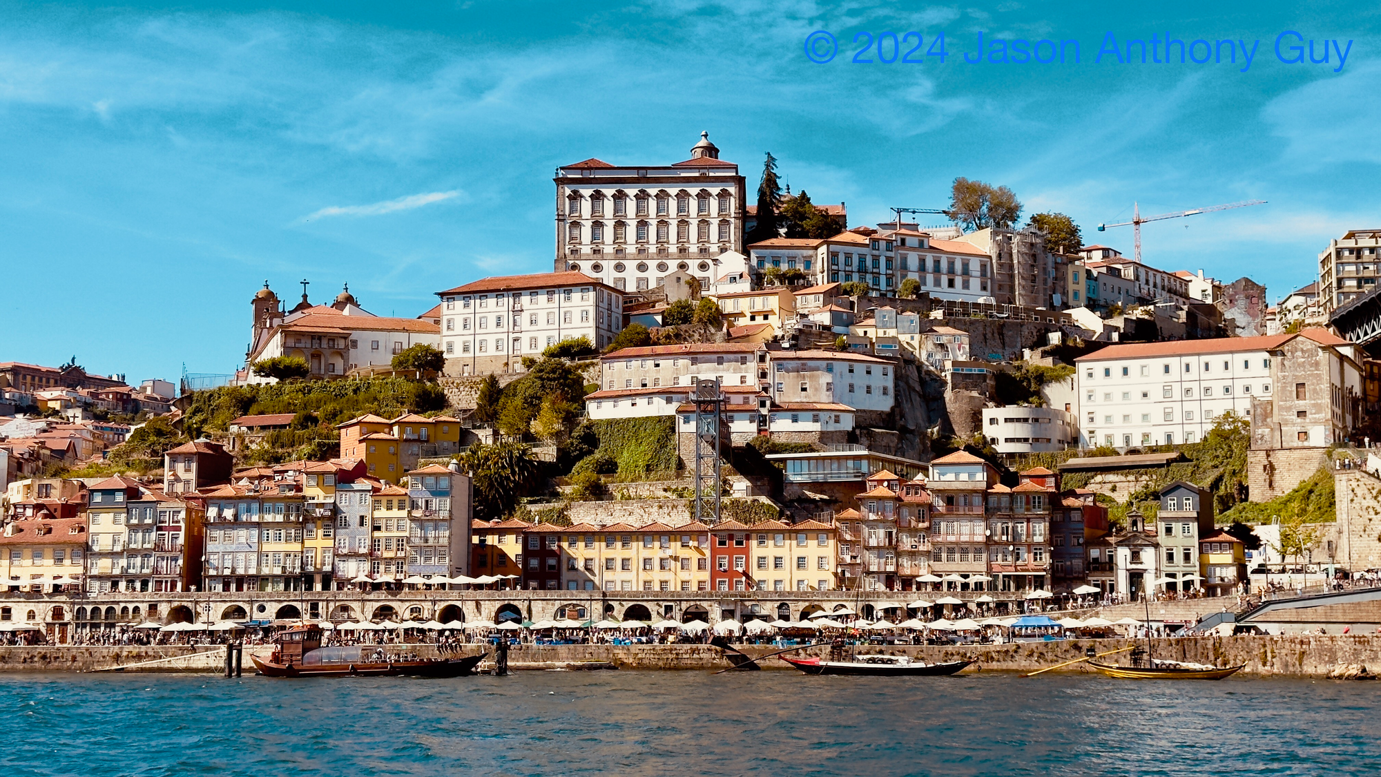 Alt: Photograph of several colorful buildings on a hill overlooking a river. The building are in yellows, reds, and creams, which contrasts with the dark blue of the river and the lighter blue of the sky.