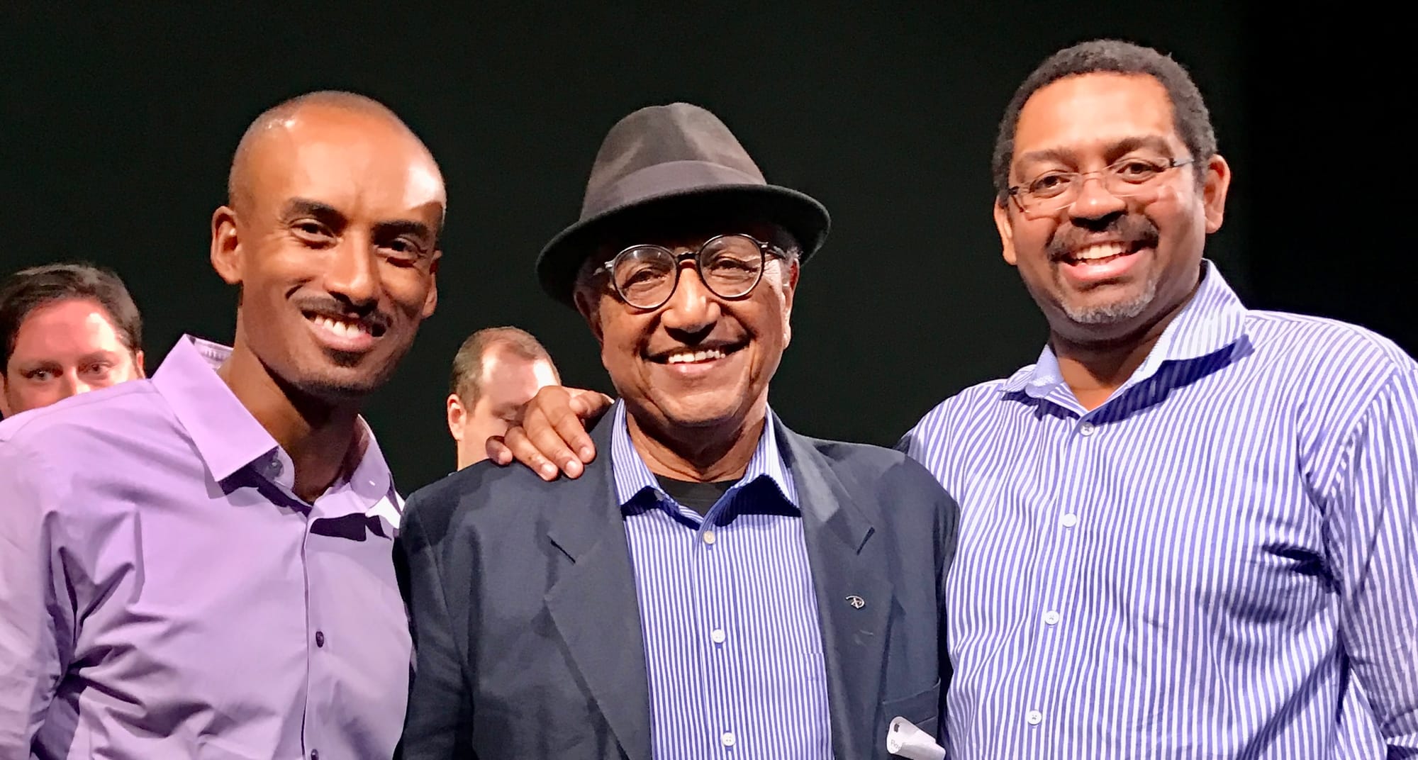 Photograph of three smiling men in collared dress shirts. The older man in the middle wears a fedora and blazer.
