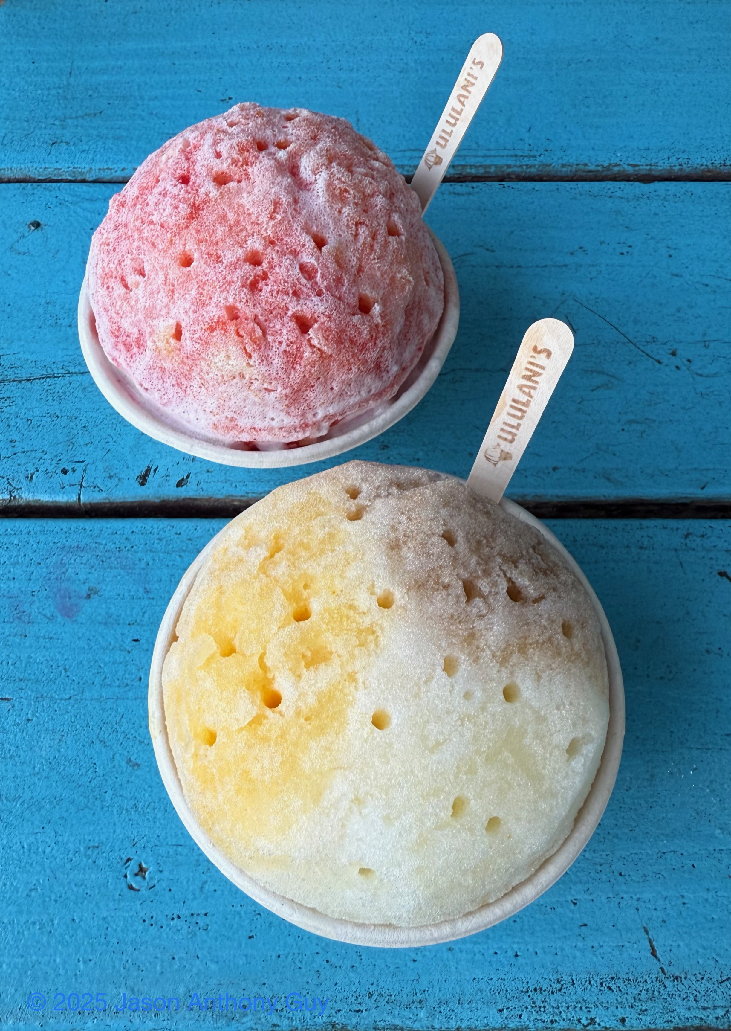 Alt: Photograph from above of two cups of shave ice, one red, one yellow and white, sitting on a blue wooden table. Both have a wooden spoon with “Ululani’s” printed on them.