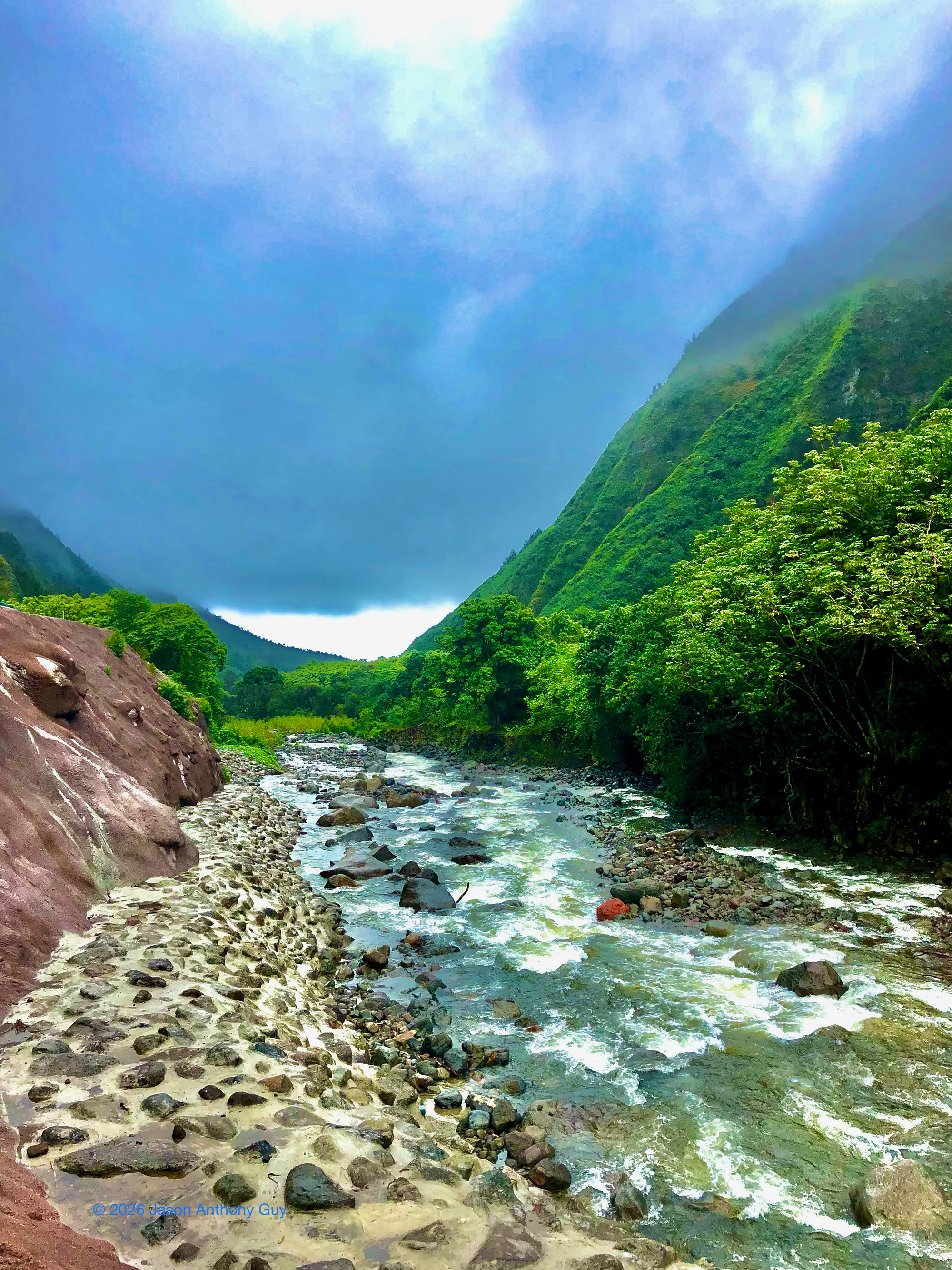 A rocky river flows through a narrow, green mountain valley. Clear water is bordered by a pebbled riverbank on the left and dense green trees on the right. Steep, forested hills rise sharply on both sides, fading into mist near their peaks. Overhead, low clouds fill the moody sky.